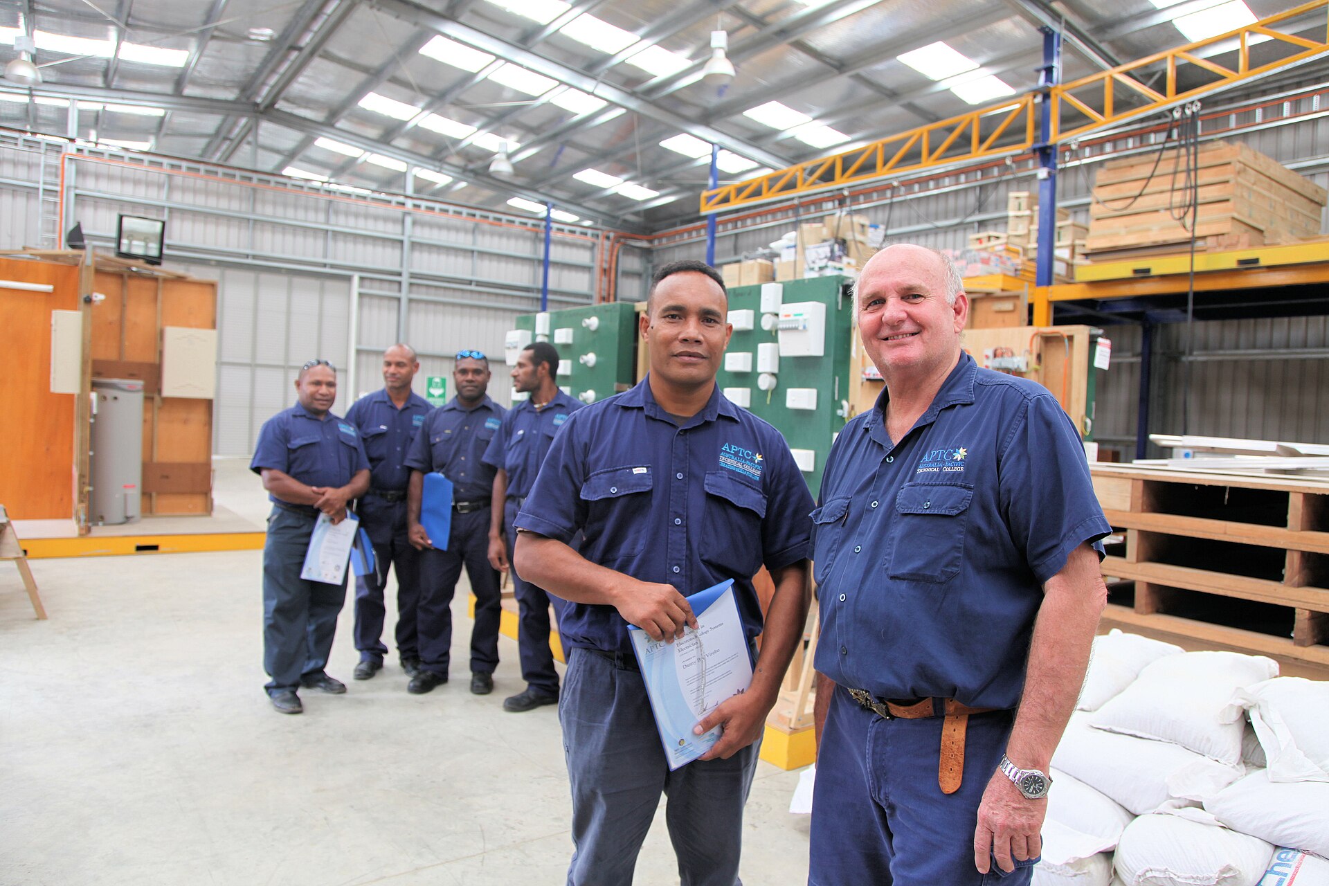 PNG electrician Danny Virobo and Australian trainer Roderick Elliot with fellow classmates in the background at the Australia-Pacific Technical College workshop on the outskirts of Port Moresby. The College is providing internationally recognised qualifications for local tradespeople in course from plumbing through to construction and hairdressing. Photo: AusAID Jacqueline Smart June 2011 Port Moresby PNG To request copyright use and access to hi-res versions of this photo, contact photolibrary@ausaid.gov.au